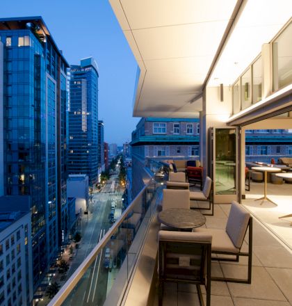 A modern balcony with tables and chairs overlooks a city street lined with tall buildings at dusk, featuring a glass railing and outdoor lighting.
