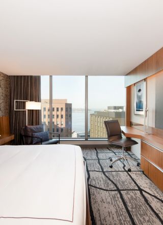 A modern hotel room with a large bed, wall-mounted TV, desk, chair, and a window offering a cityscape view.
