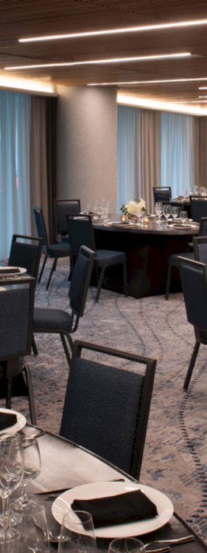 An elegant banquet hall with round tables set for an event, featuring black chairs, table settings, floral centerpieces, candles, and modern lighting.