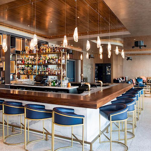 A modern bar with blue chairs, a wood counter, and pendant lights. Shelves behind the bar are stocked with various bottles.