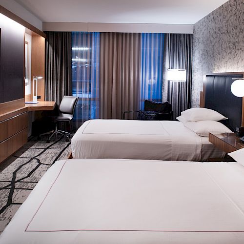 This image shows a modern hotel room with two beds, a wall-mounted TV, a desk and chair near the window, and stylish decor.