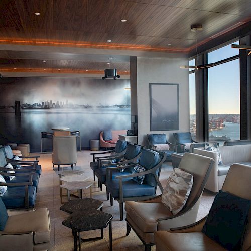 The image shows a modern lounge with comfortable seating, large windows offering views of a waterfront and a ferris wheel, and a serene ambiance.
