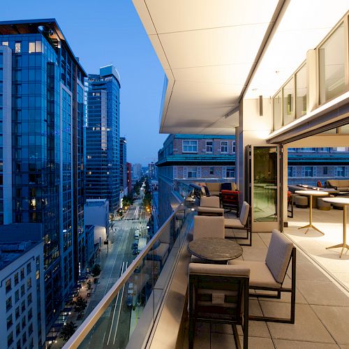 A modern balcony at dusk overlooks a city street lined with tall buildings. The balcony has chairs, tables, and indoor-outdoor seating areas.