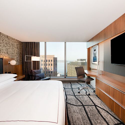 A modern hotel room with a large bed, a desk and chair, a flat-screen TV, and a view of buildings and water through large windows.