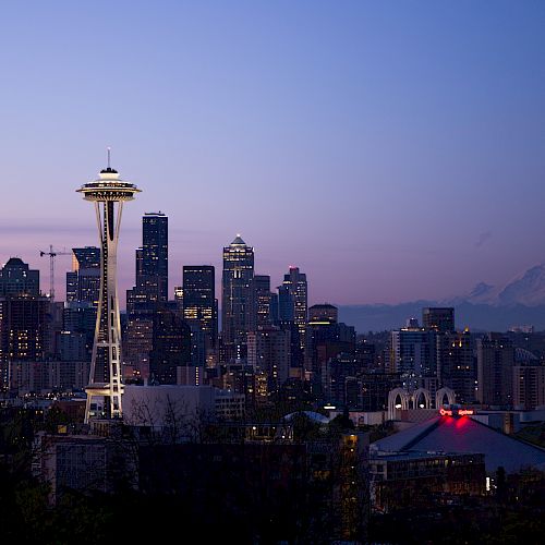 A cityscape of Seattle at dusk featuring the iconic Space Needle with Mount Rainier in the background and buildings illuminated in the twilight.