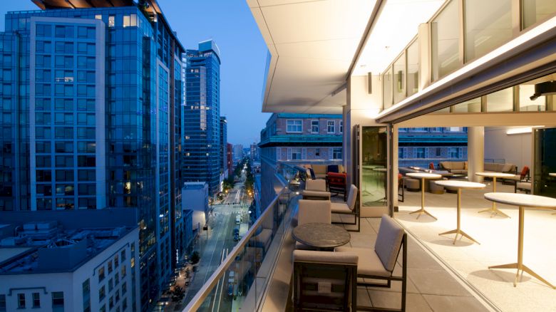 A modern rooftop terrace with seating overlooks a cityscape at dusk, featuring tall buildings and a mix of commercial and residential spaces.