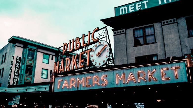 An image of a public market with neon signs reading "PUBLIC MARKET" and "FARMERS MARKET" below a clock, with surrounding buildings visible.