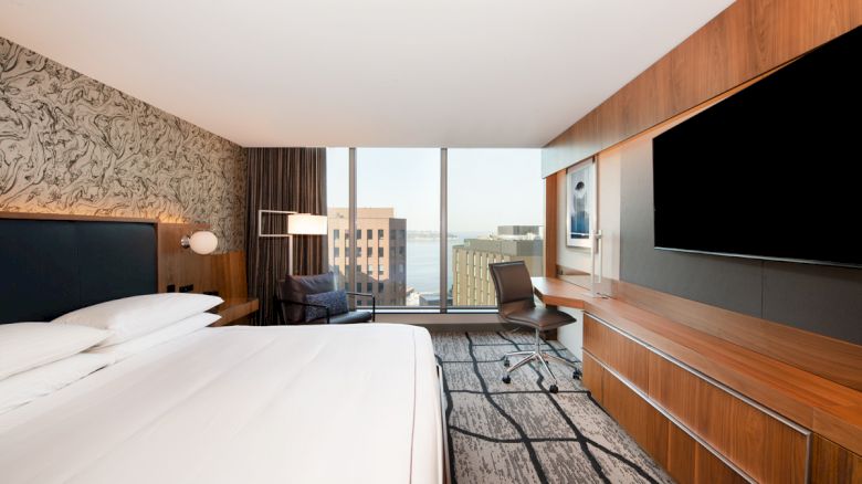 A modern hotel room with a large bed, desk, chair, wall-mounted TV, and a window with a city view and partial ocean view in the background.