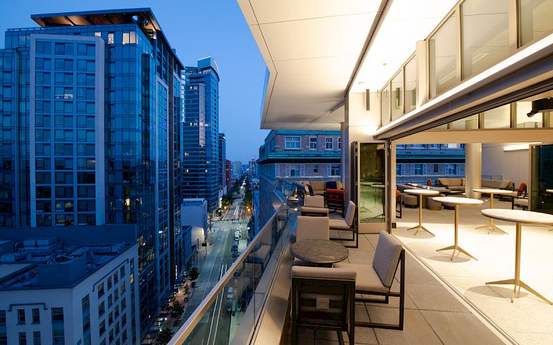 A modern balcony with seating overlooks a cityscape of high-rise buildings under a dusky blue sky, showcasing urban architecture and evening lights.