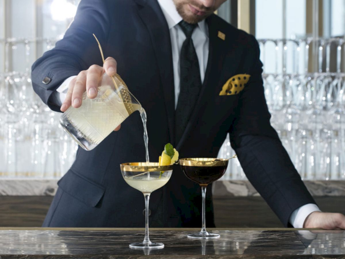 A person in a suit is pouring a drink from a glass into a cocktail glass garnished with lemon and a mint leaf beside another cocktail glass.