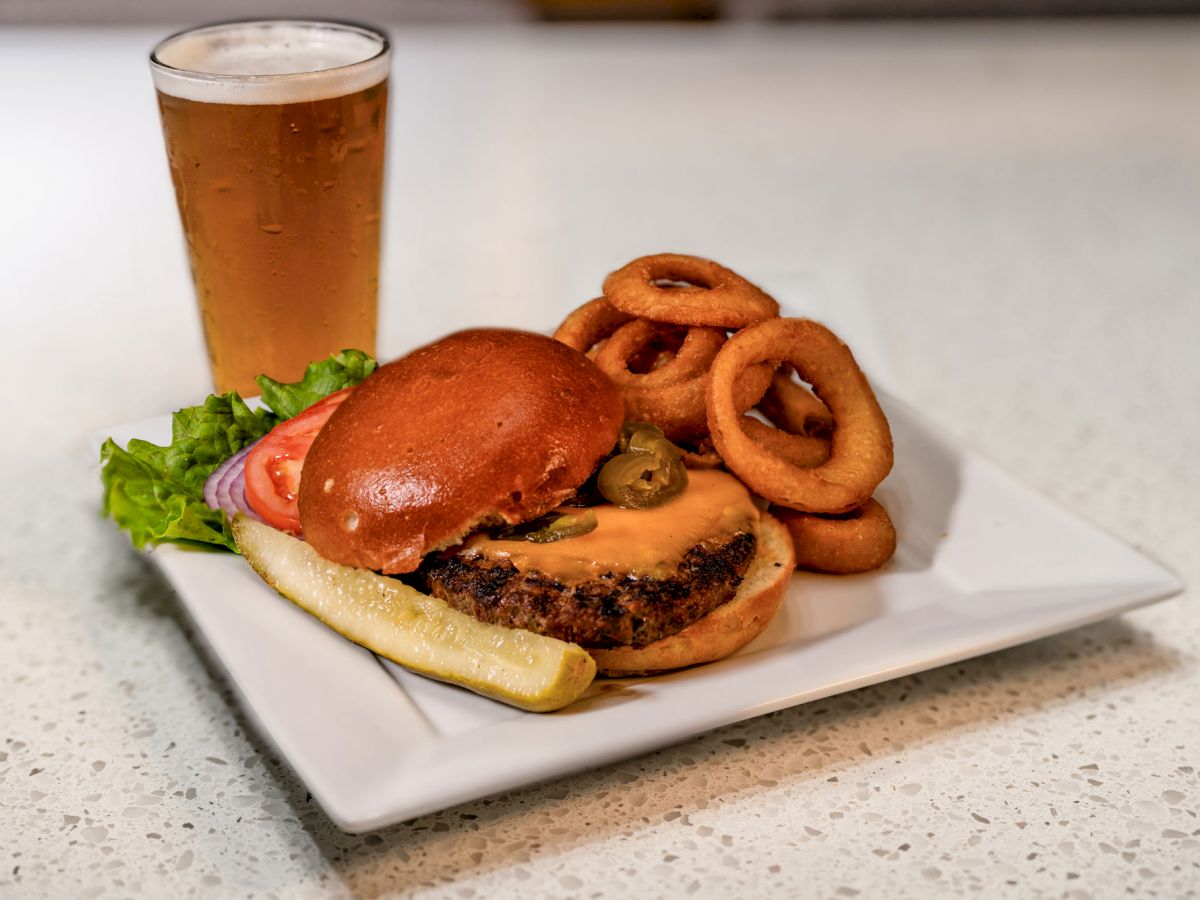 The image shows a plate with a cheeseburger, onion rings, a pickle, and a side of lettuce and tomato, along with a glass of beer.