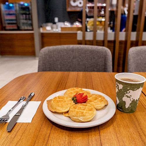 A dining setup with a plate of waffles topped with a strawberry and a cup of coffee on a wooden table, along with cutlery and a napkin.